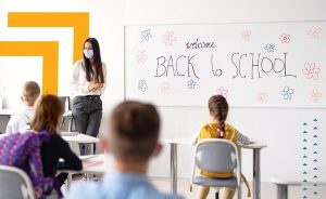 Teacher wearing mask and students in a classroom with a 'Back to School' message on whiteboard.
