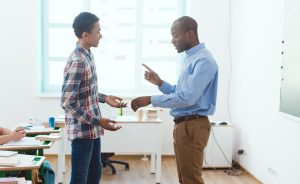 Male teacher in a blue shirt and khaki pants talks with a young male student in a classroom. The teacher is gesturing with his hands.
