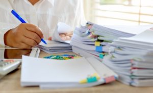 A desk covered in stacks of papers being graded, with paperclips and a teacher writing with a blue pen.