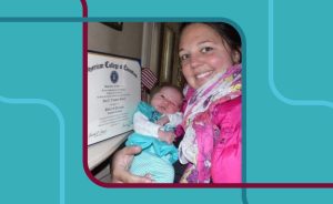 A woman with dark hair smiles and holds a newborn baby in her arms. A framed certificate is visible in the background.