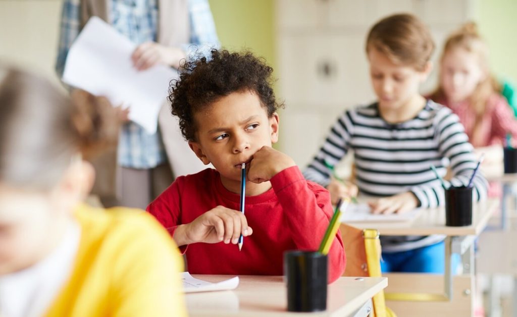 Elementary school classroom scene with a young boy in the foreground deep in thought with a pencil. Other children are seated at desks around him, and a teacher is standing in the background.