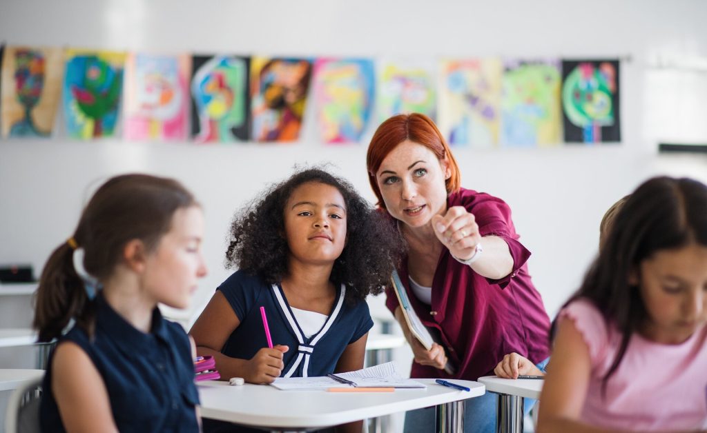 Female teacher leans over a young female student who is working at a desk in a classroom with other students and colorful artwork on the wall.