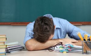 Male teacher resting head on desk with papers.