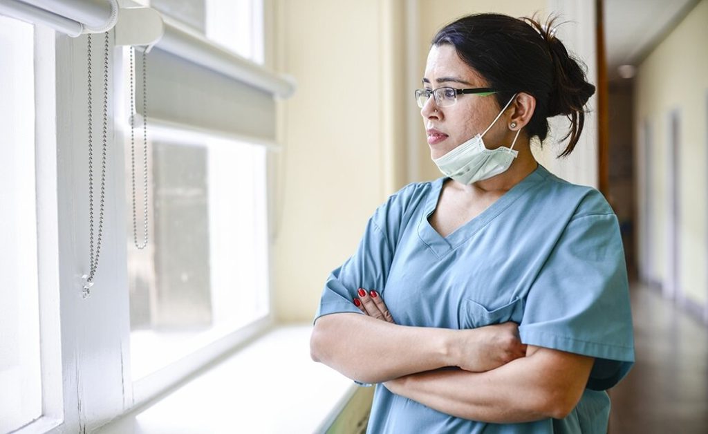 Female nurse wearing scrubs and a face mask looking out a window of the hospital with arms crossed.