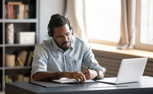 A young Black man wearing headphones and glasses is writing in a notebook while using a laptop.