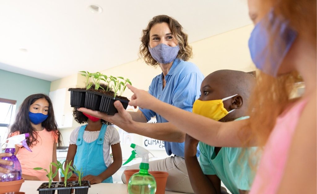 A teacher wearing a face mask shows seedlings to students, who are also wearing masks