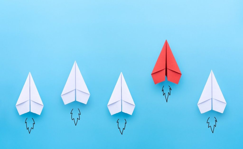 A visual representation of leadership, standing out, or being different. Four white paper airplanes follow a slightly leading and higher red paper airplane against a blue backdrop.