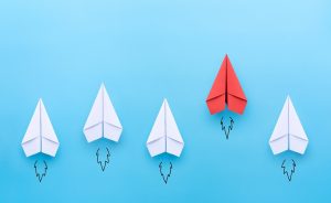 A visual representation of leadership, standing out, or being different. Four white paper airplanes follow a slightly leading and higher red paper airplane against a blue backdrop.