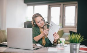 Female teacher with headset sits at desk with laptop, tablet, and coffee, holding up a handwritten list and pointing to it.