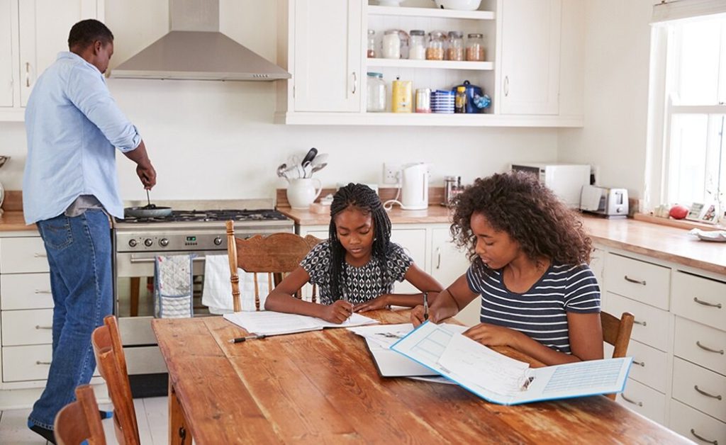 A father cooks in the kitchen while two daughters do homework at a table.