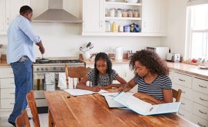 A father cooks in the kitchen while two daughters do homework at a table.