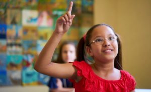 Young girl wearing glasses and a red shirt with her hand raised in a classroom.