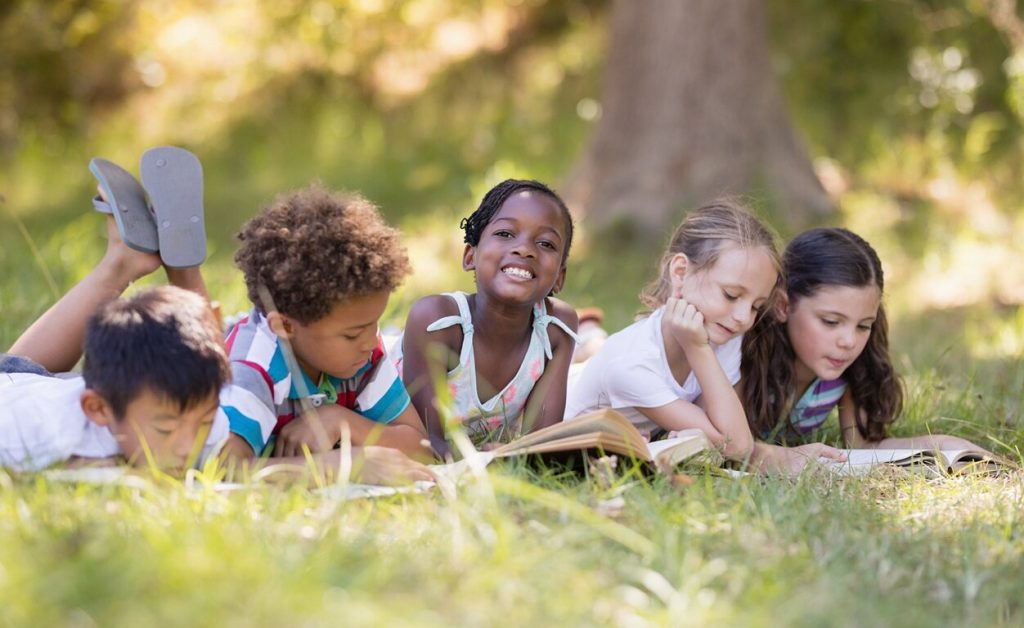 Children reading books outdoors on grass.