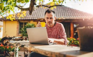 A man sits outdoors working on a laptop and holding a yellow mug.