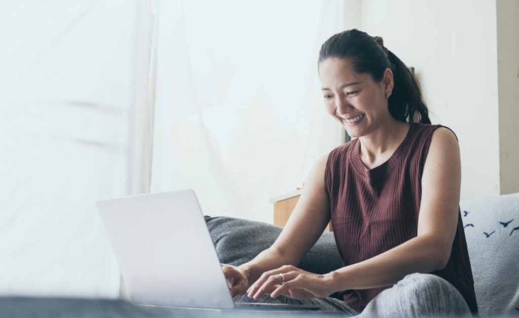 A woman smiles while using a laptop on a couch.