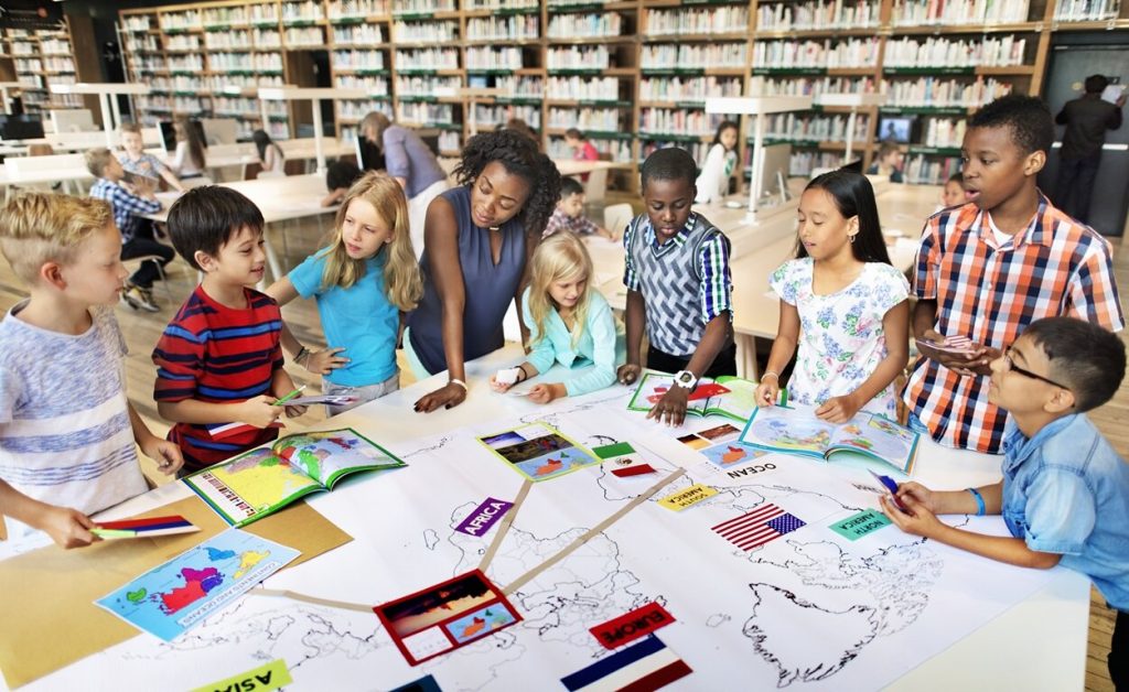 Female teacher with young students looking at map on a table in the library.