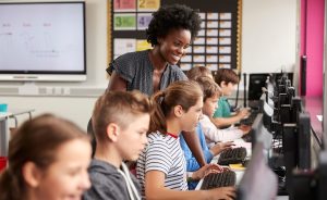 Female teacher assisting elementary school students working on computers in a classroom.