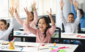 Enthusiastic elementary school students raise their hands in a classroom.