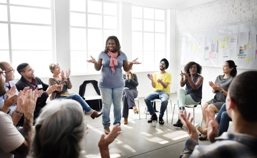 A person in a leadership role is speaking to a group of people who are clapping during a meeting.