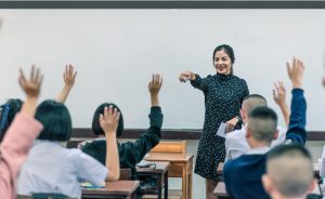 Female teacher interacts with students raising their hands, possibly at the start of a new school year.
