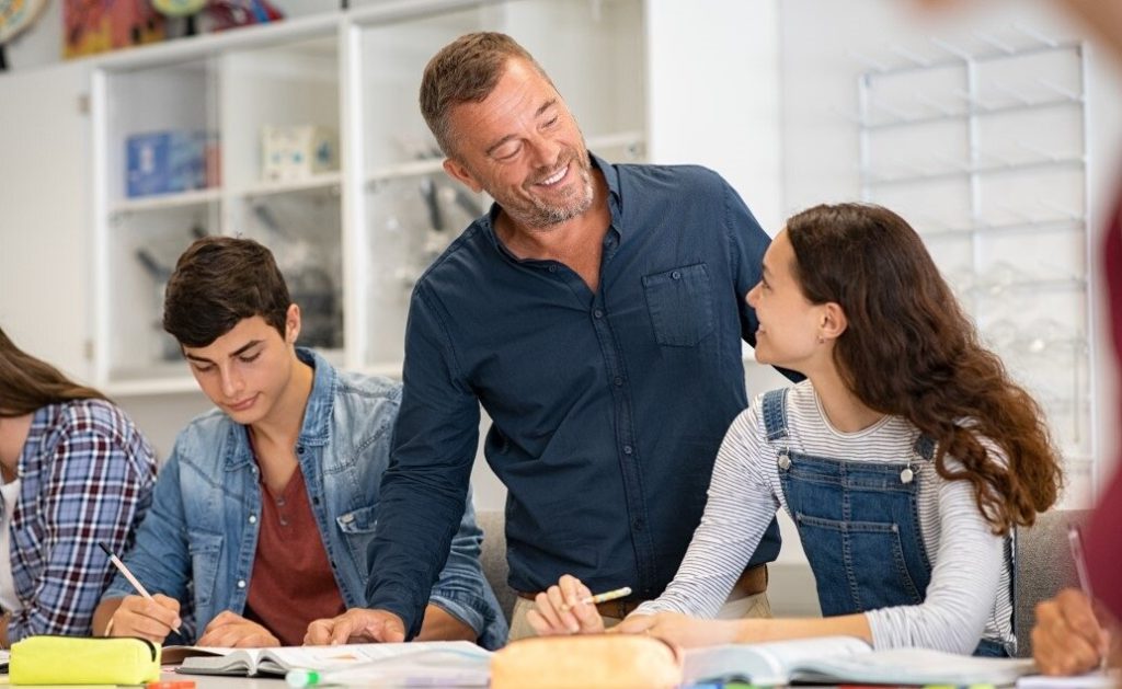Male teacher helping student while other students are seated at the same table, working on assignments.
