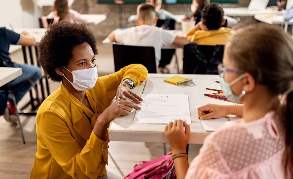 A teacher wearing a face mask appears to be reviewing a test with a student who is also masked.