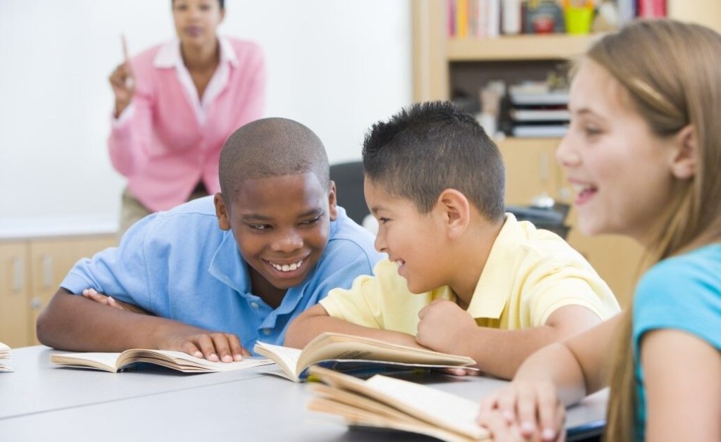 A group of students are talking at desks while a female teacher is teaching in background.