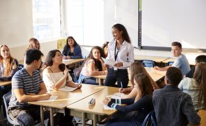 Female teacher addressing a group of high school students sitting at desks in a classroom.