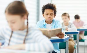 A young Black student reading a book in a classroom with other students.