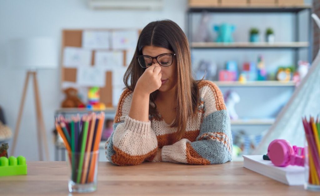 A female teacher with glasses looks stressed at a desk.