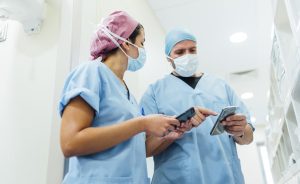 Two nurses in a hopsital wearing scrubs and face masks looking at mobile phones together