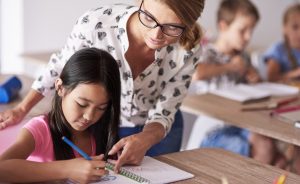 Female teacher helps young girl writing in notebook at desk in classroom with other students.