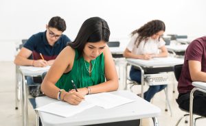 Four high school students sit at individual desks in a classroom, focused on writing on papers in front of them.