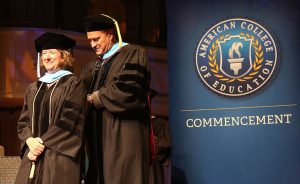 Graduation ceremony with a graduate and faculty member in front of an American College of Education banner