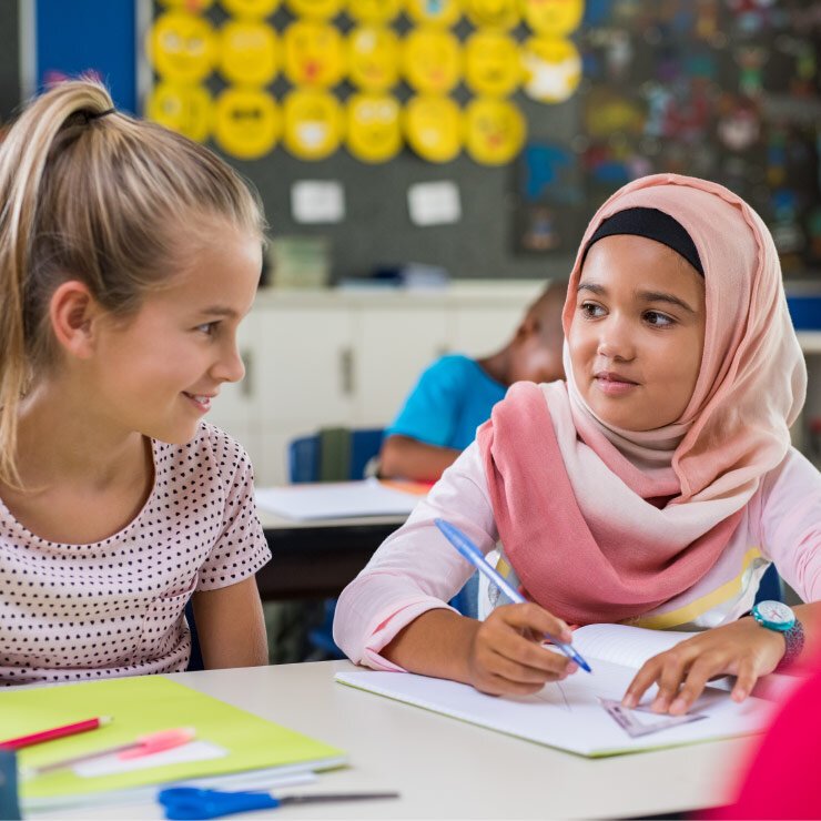 Two young girls talking while sitting together at a table at school. One is wearing a hijab and writing in a notebook.