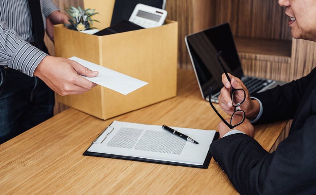 Photo of a man handing a white envelope to another man at a desk, with a cardboard box and laptop in the background, possibly depicting a resignation.