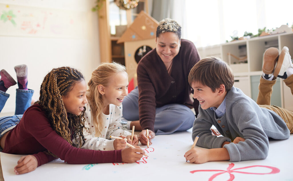 Female teacher coloring with three young students on large paper on the ground