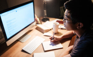 Male student working on computer while writing on a notepad.