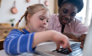 Female African American teacher helping a young student with a learning disability type on a laptop.