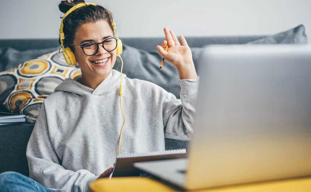 Young female student with yellow headphones on waving at laptop.