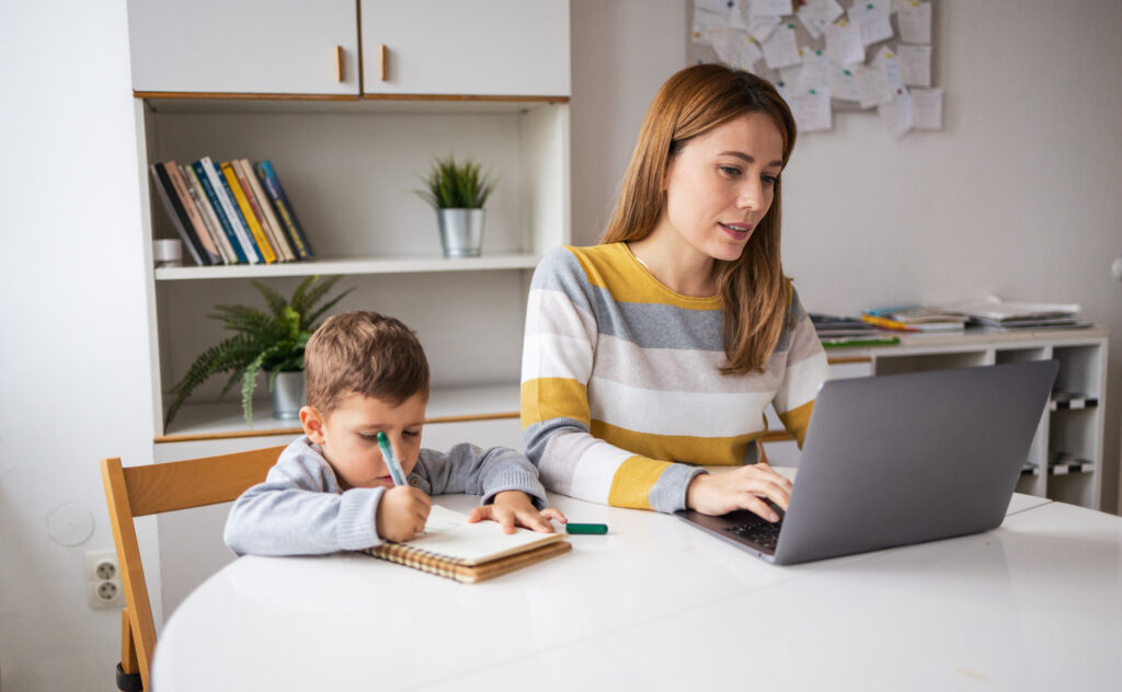 Mother working on homework on laptop while child next to her draws on a notebook.