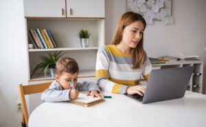 Mother working on homework on laptop while child next to her draws on a notebook.