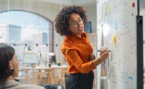 Black woman in professional clothing stands in front of a whiteboard with marker in hand, writing as she speaks