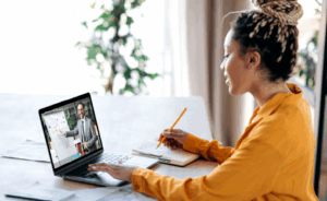 Woman sitting at a table, watching an instructional video on her laptop while she takes notes in a notebook with a pencil